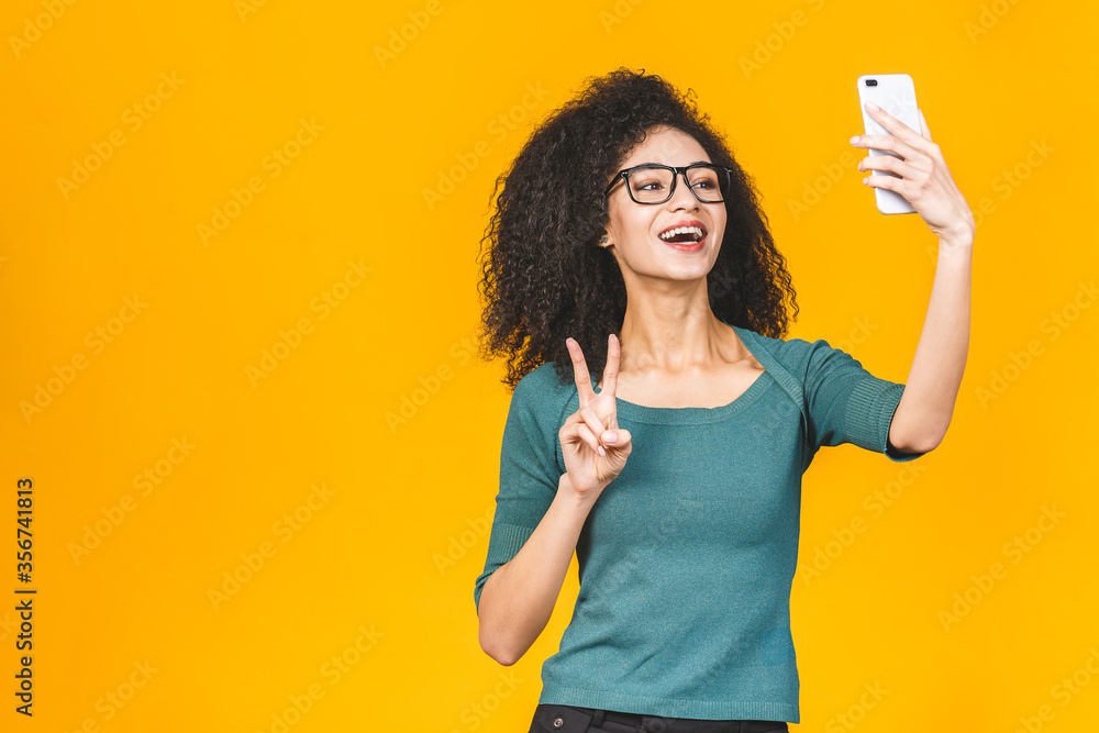 Young funny african american casual lady grimacing and making selfie on smartphone isolated over yellow background. Young funny african american casual lady grimacing and making selfie on smartphone isolated over yellow background.