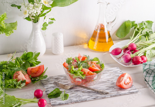 Salad with vegetables and herbs in a transparent salad bowl on a white wooden background.