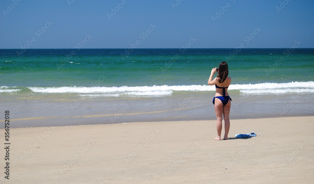 Menina parada na praia em biquini azul a fotografar a paisagem marítima com o telemóvel, calções pousados no chão ao lado, momento de descontracção Menina parada na praia em biquini azul a fotografar a paisagem marítima com o telemóvel, calções pousados no chão ao lado, momento de descontracção