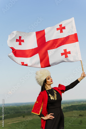 Georgian girl holds national flag of Georgia on blue sky background. Georgian culture lifestyle. Woman in papakha and red dress photo