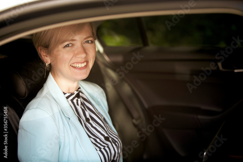 happy young woman in a blue jacket in business clothes sitting in the back seat of a car with a laptop