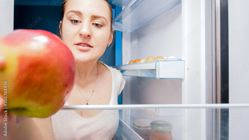Portrait of hungry woman taking red apple out of fridge on kitchen at night. Concept of dieting and healthy nutrition Portrait of hungry woman taking red apple out of fridge on kitchen at night. Concept of dieting and healthy nutrition