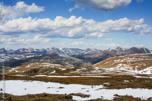 View of Trail Ridge Road traveling through Rocky Mountain National Park with snowy peaks of "Never Summer" mountains in distance, blue cloudy sky and snow covered foreground