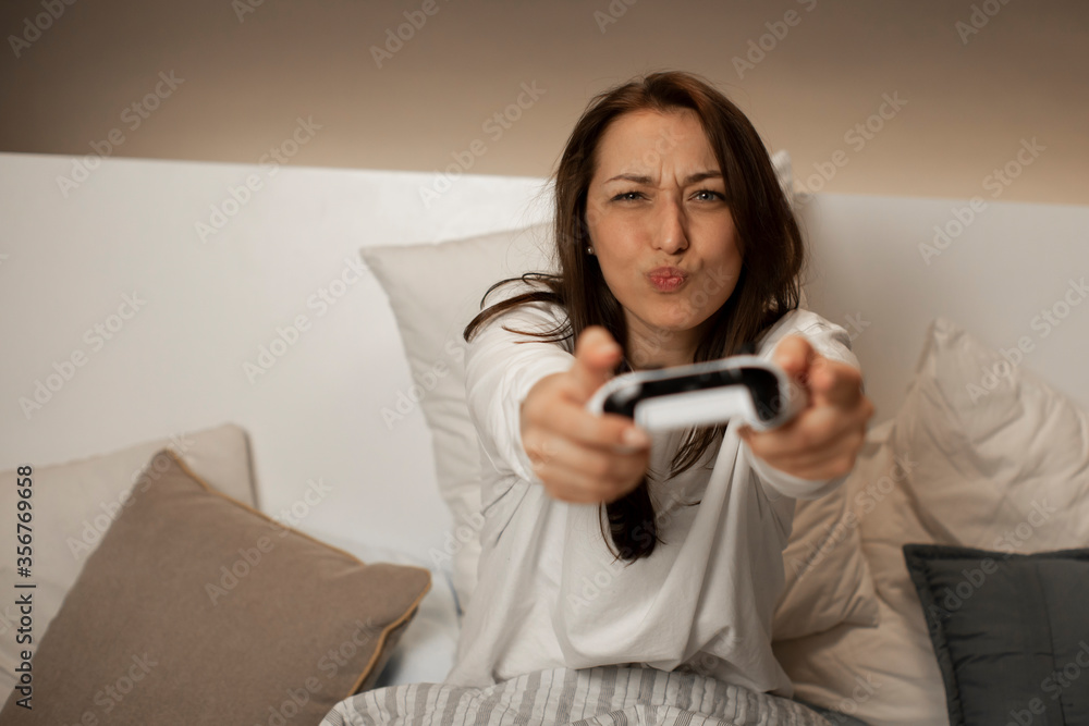 Caucasian young female with dark hair in white pajamas in bedroom with stylish lighting plays video games on the console using the joystick, smile and make antics. Quarantine entertainment concept. Caucasian young female with dark hair in white pajamas in bedroom with stylish lighting plays video games on the console using the joystick, smile and make antics. Quarantine entertainment concept.