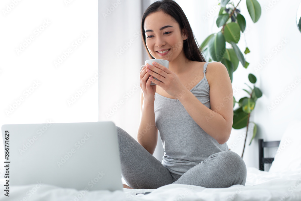 Happy woman with coffee looking at laptop screen in bedroom Happy woman with coffee looking at laptop screen in bedroom