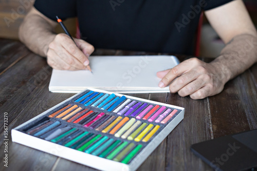 person, artist painting with a pastel crayon chalk on sketchbook on the wooden table, selective focus.