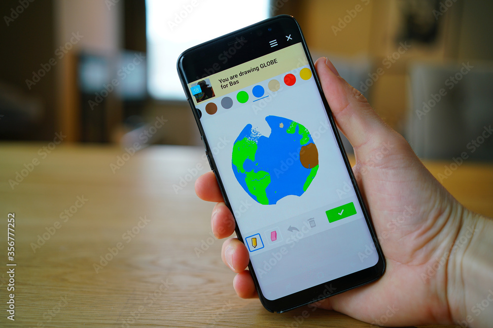 A hand holding a modern black smartphone with a generic drawing game on the touch screen. The current drawing in the fun application is a globe. Games on phones are becoming increasingly popular.