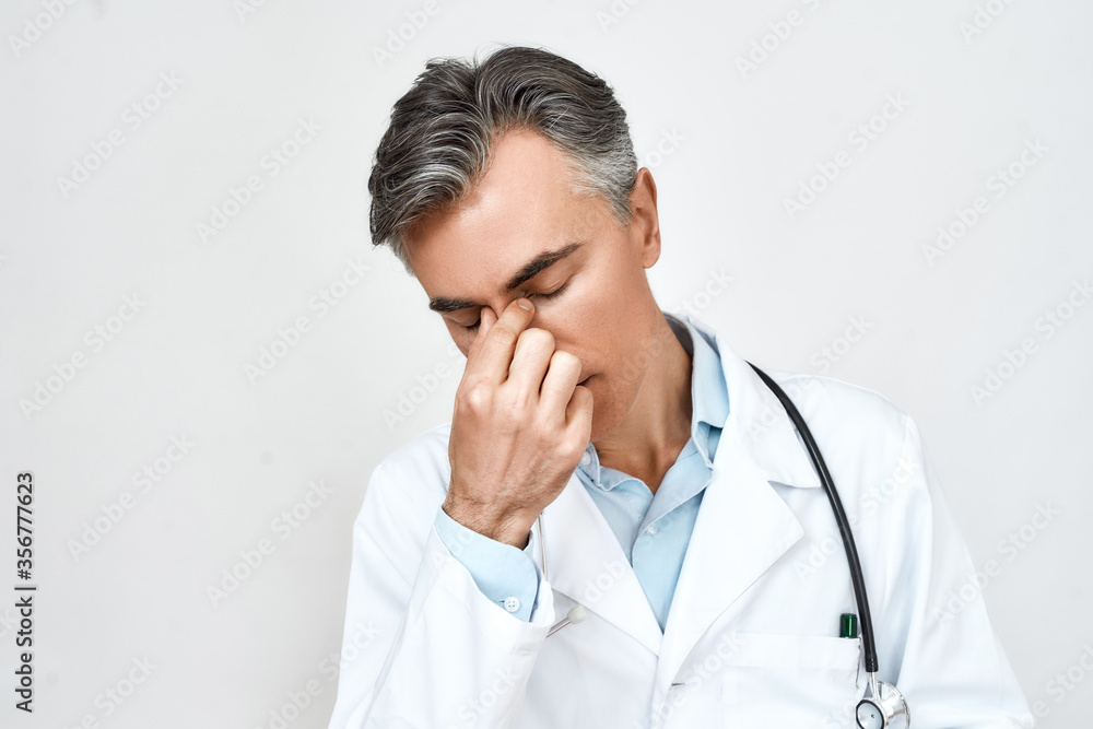 Portrait of tired stressed male doctor in medical uniform feeling upset, keeping eyes closed and touching nose while standing against grey background Portrait of tired stressed male doctor in medical uniform feeling upset, keeping eyes closed and touching nose while standing against grey background