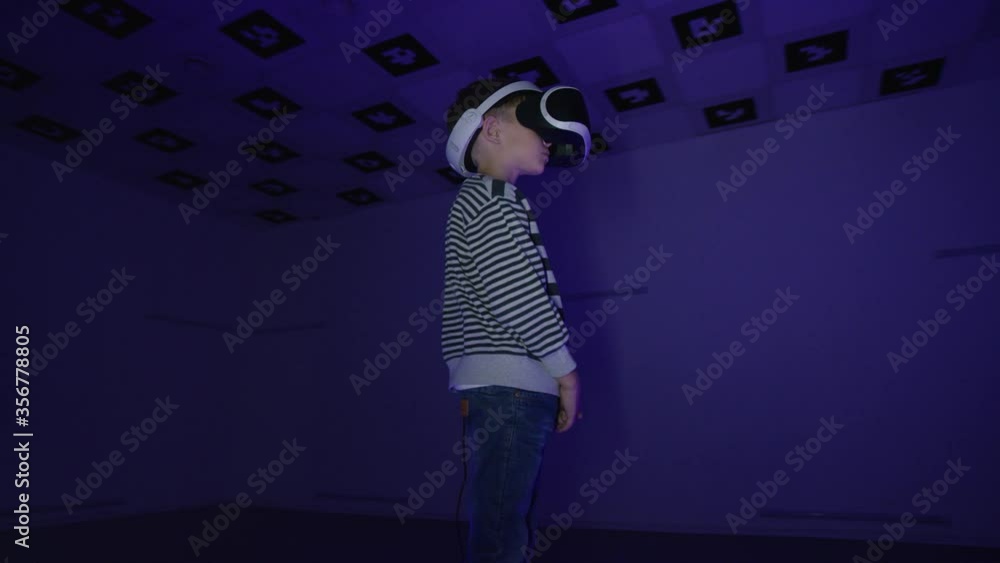 Tracking arc 360 degree shot of a little boy is looking around in virtual reality wearing headset in a room with flickering red and blue neon lights Tracking arc 360 degree shot of a little boy is looking around in virtual reality wearing headset in a room with flickering red and blue neon lights