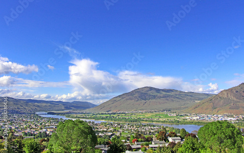 Kamloops, BC. The aerial view on the Thompson river and the city surrouned by hills / moutains from the look out point. Green trees in the foreground, blue sky in the background. © Klara