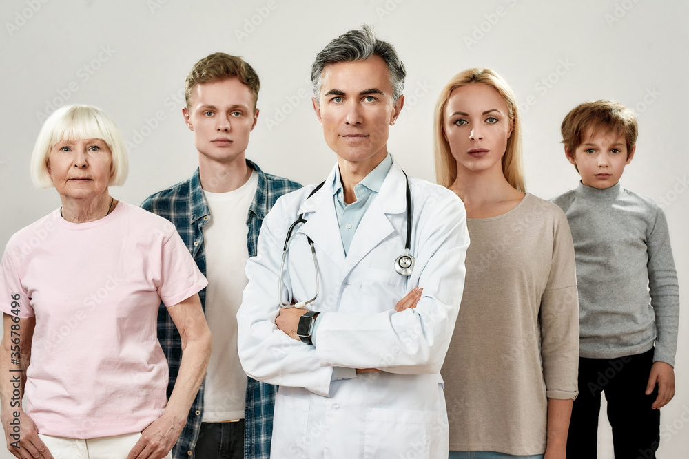 Portrait of professional mature male doctor in medical uniform and family of four people looking at camera while standing against grey background Portrait of professional mature male doctor in medical uniform and family of four people looking at camera while standing against grey background