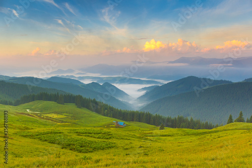Magic dawn on a pasture in the mountains. Shepherd's house in the meadow. The fog spreads in the valley. Beautiful clouds in the blue sky. Ukraine, Europe.