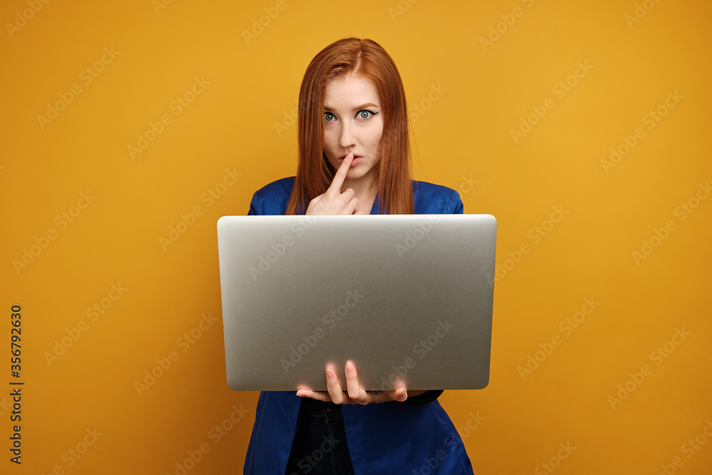 Naklejka premium A red-haired girl in a blue jacket stands on a yellow background with a laptop in hand and putting finger to lips