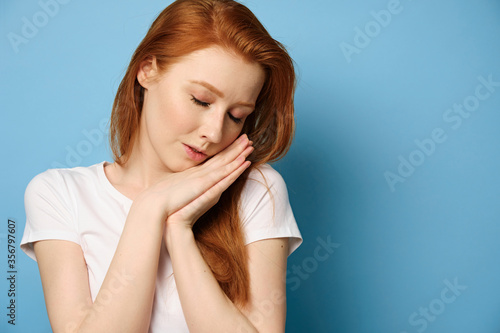 Red-haired girl in a white T-shirt stands on a blue background, resting her head on folded palms and closing her eyes.