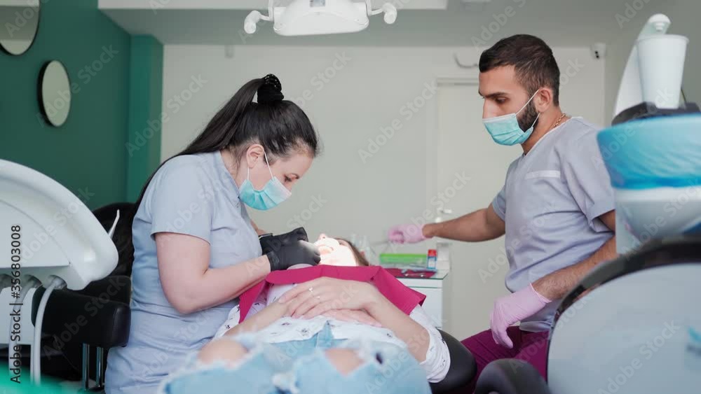 Two Doctors Dentists Woman and Man In Medical Masks Perform Surgery to Patient Girl in the Dental Office. Healthcare and medicine concept Two Doctors Dentists Woman and Man In Medical Masks Perform Surgery to Patient Girl in the Dental Office. Healthcare and medicine concept