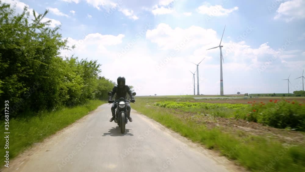 Motorcyclist in black rides on a path between fields with wind turbines in the background on a sunny day - Front view tracking shot Motorcyclist in black rides on a path between fields with wind turbines in the background on a sunny day - Front view tracking shot