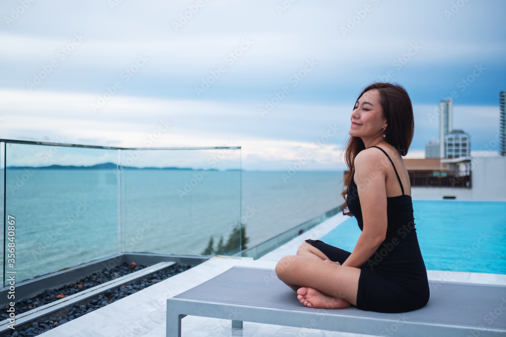 A beautiful young asian woman sitting next to the rooftop pool with sea and blue sky view