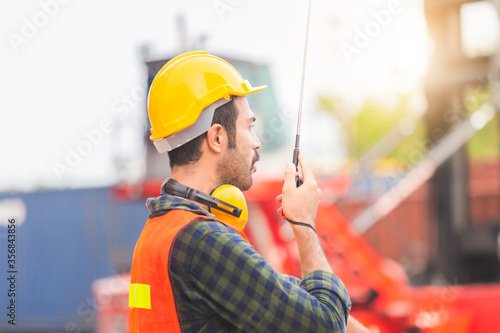 Foreman worker in hardhat and safety vest talks on two-way radio control loading containers box from cargo
