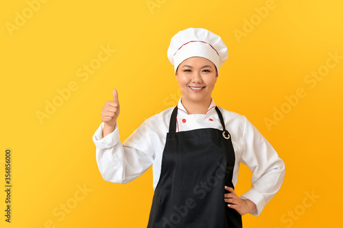 Beautiful Asian chef showing thumb-up gesture on color background