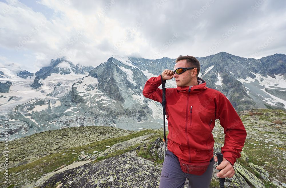 Close up of tourist taking on eyeglasses, walking using trekking sticks, beautiful mountains with snow on background. Mountain hiking, man reaching peak. Sport tourism in Alps.
