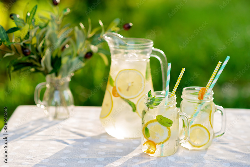 Fototapeta premium Lemonade refreshing drink in a jug and jars with lemons, fresh mint and ice on a garden table. Summer picnic outdoors. Copy space. Soft selective focus.