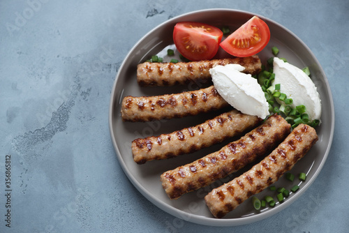 Round grey plate with grilled serbian cevapi or cevapcici sausages and kajmak cheese, studio shot on a grey concrete background with space