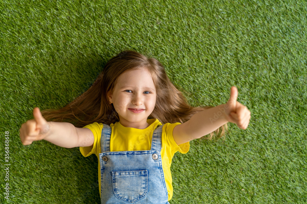 Naklejka premium cheerful little girl in a denim jumpsuit, lies on a green carpet, lawn.