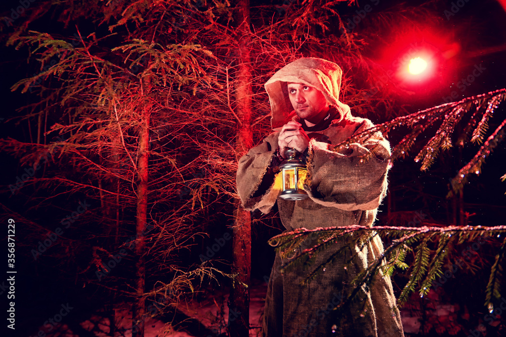 Obraz premium Medieval monk in canvas sackcloth robe with lattern in dark forest with snow and red light on winter night. Fantasy or fairy tale about wandering monk. Story about the forces of good and evil in world