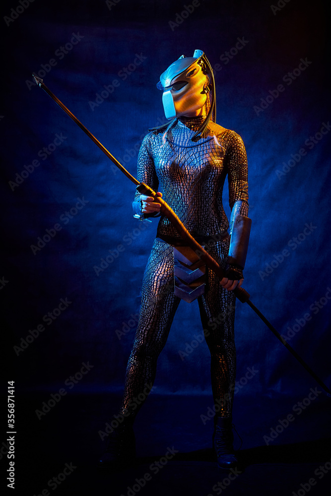 Naklejka premium Valkyrie girl looking like an alien in shiny military armor, a strange helmet and a spear in a dark room with plants and vines. Model during a photo shoot, the actress during the shooting of the film.