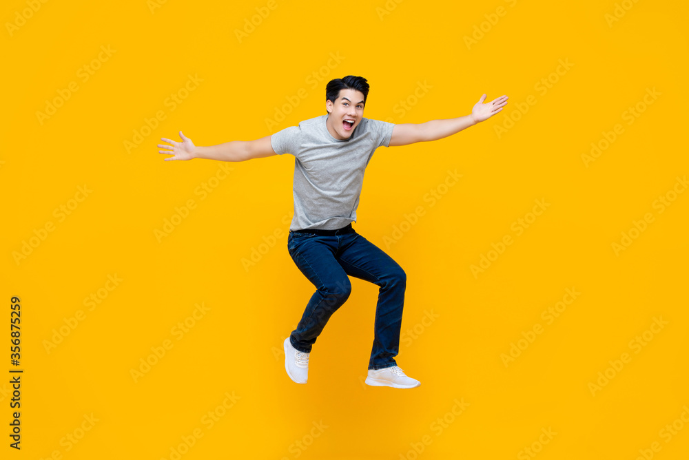 Young cheerful energetic handsome Asian man jumping with open hand gesture isolated on yellow studio background