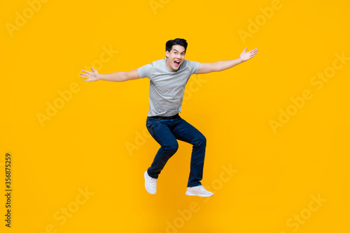 Young cheerful energetic handsome Asian man jumping with open hand gesture isolated on yellow studio background