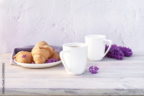 Two beautiful porcelain cups of coffee with milk with croissants decorated with lilac flowers on white wooden table. Perfect breakfast concept. Copy space.