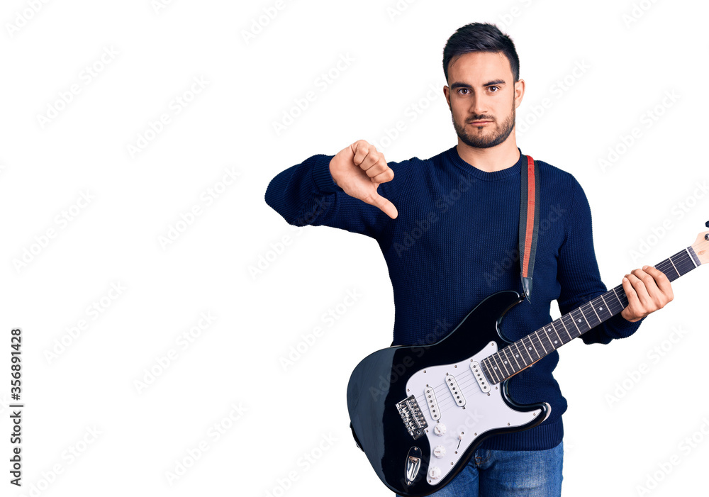 Young handsome man playing electric guitar with angry face, negative sign showing dislike with thumbs down, rejection concept Young handsome man playing electric guitar with angry face, negative sign showing dislike with thumbs down, rejection concept