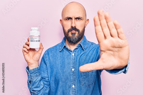 Young handsome man holding pills with open hand doing stop sign with serious and confident expression, defense gesture