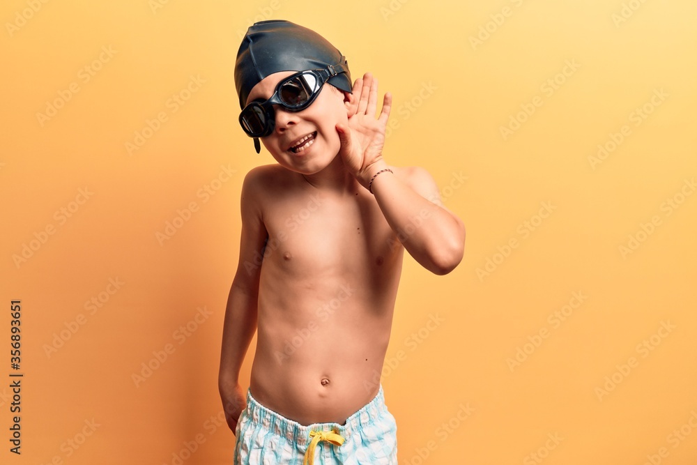 Cute blond kid wearing swimwear and swimmer glasses smiling with hand over ear listening and hearing to rumor or gossip. deafness concept.