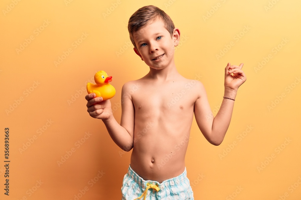 Cute blond kid wearing swimwear holding duck toy smiling happy pointing with hand and finger to the side Cute blond kid wearing swimwear holding duck toy smiling happy pointing with hand and finger to the side