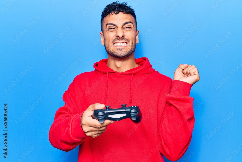 Young hispanic man playing video game holding controller screaming proud, celebrating victory and success very excited with raised arm Young hispanic man playing video game holding controller screaming proud, celebrating victory and success very excited with raised arm