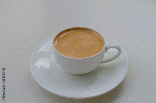 A small white cup with aromatic light brown coffee on a saucer, on a light background