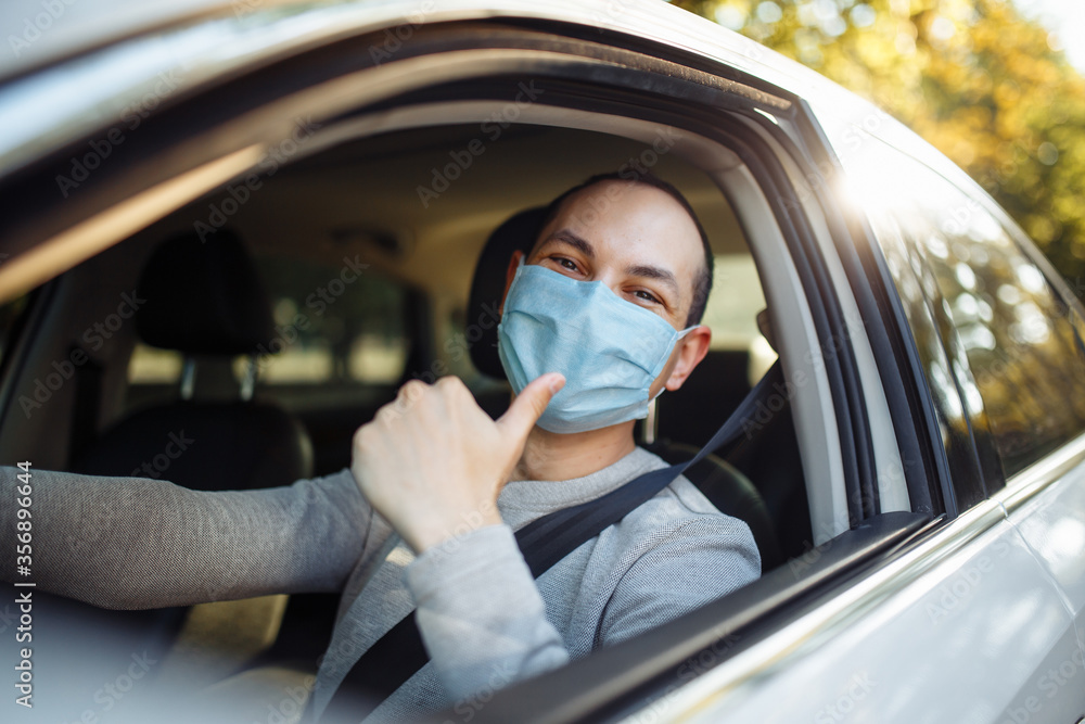 Young man shows thumb up like sign wearing sterile mask in the car. Taxi driver delivers his passanger to the destination. Social distance, new normal, virus spread prevention and treat concept. Young man shows thumb up like sign wearing sterile mask in the car. Taxi driver delivers his passanger to the destination. Social distance, new normal, virus spread prevention and treat concept.