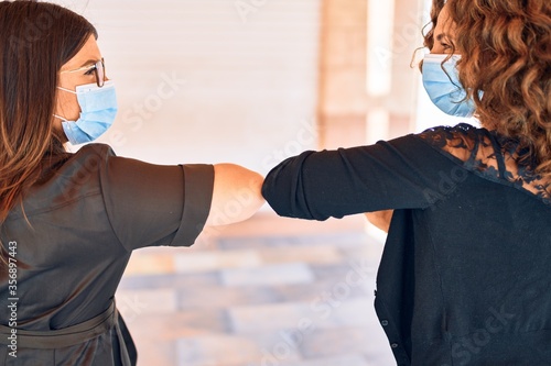 Two beautiful friends doing new elbow greeting to coronavirus prevention. Wearing medical mask standing at town street. photo