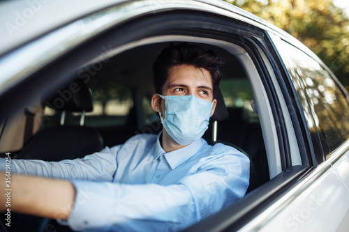 Young man sits behind the steering wheel in the car wearing sterile medical mask. Boy taxi driver works hard during coronavirus outbreak. Social distance, virus spread prevention and treat concept.