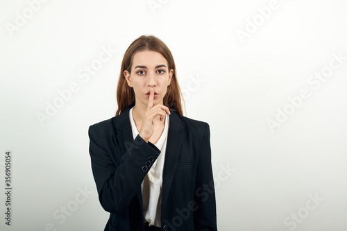 Index finger on lips, silence gesture, shhh quiet, asks for voicelessness forefinger at the mouth. Young attractive woman with brown hair in a light t-shirt and black jacket on white background