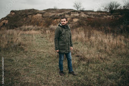 Handsome young stylish man in a green jacket, Scandinavian Christmas sweater and jeans walks in nature. Cloudy weather, spring, dry grass.
