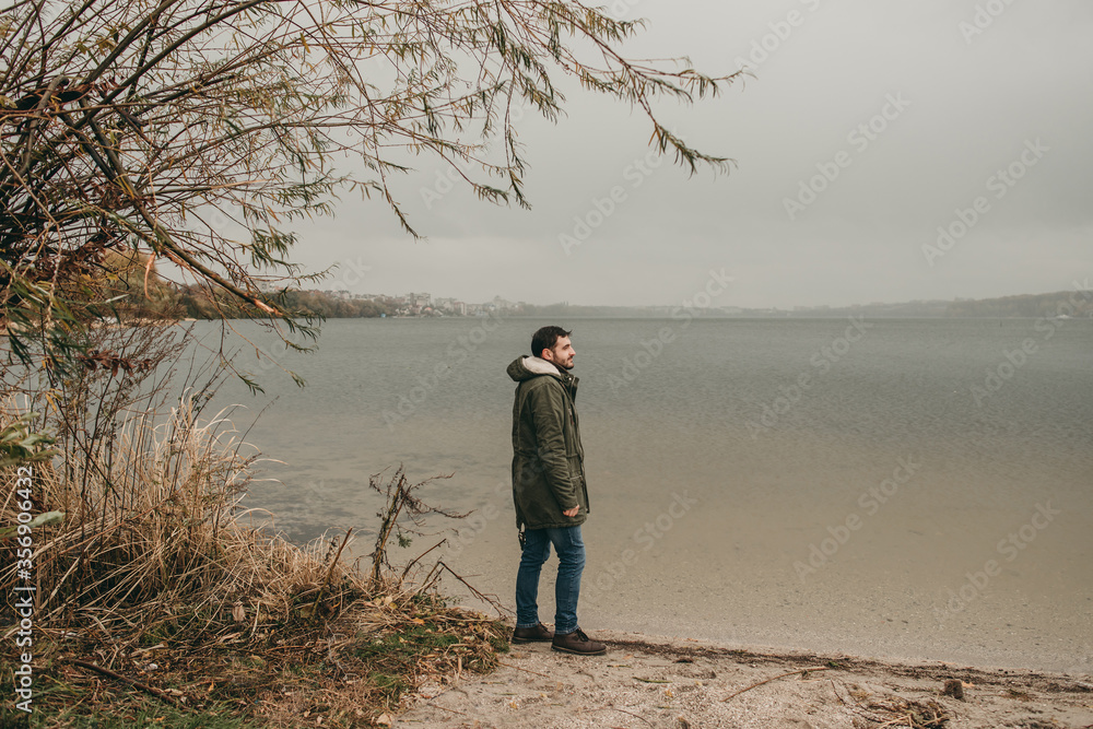 Young sad man in a green jacket walks in nature near the lake in autumn. Traveling, cold weather, rain, thought, thoughtfulness and loneliness.	