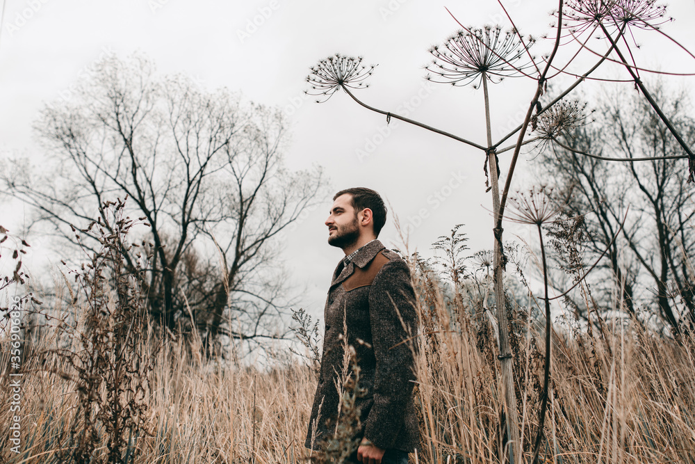 Handsome sadly young stylish man in a jacket and jeans walks on the nature of jubilee dried autumn flowers. Cloudy weather, spring, dry grass Handsome sadly young stylish man in a jacket and jeans walks on the nature of jubilee dried autumn flowers. Cloudy weather, spring, dry grass