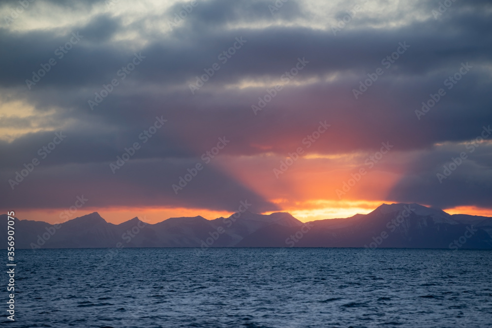 Sunset during the Arctic summer on Spitsbergen. Warm light with pink sun beams going up in the sky. Snow covered mountains at the horizon.