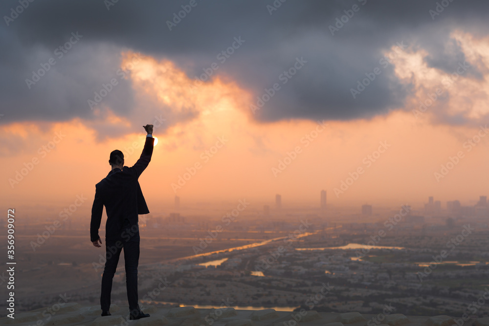 Victory and success concept. Young male businessperson with hand in air and thumbs up standing confident on top of high-rise building with beautiful sunrise or sunset view in the background.
