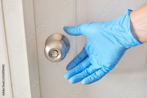 Hand with blue glove going to hold the doorknobs of the closed white door. Health concept to aware for touching the everyday objects.