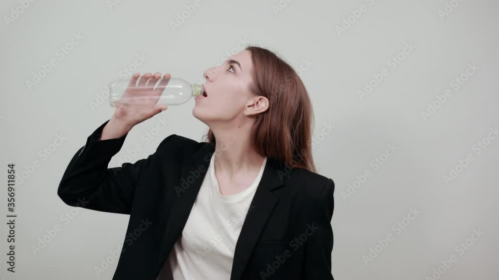 Drinking water from plastic bottles, quenches thirst after fitness indoors. Lose weight. Healthy lifestyle. Young attractive woman with brown hair in a light t-shirt, black jacket on white background Drinking water from plastic bottles, quenches thirst after fitness indoors. Lose weight. Healthy lifestyle. Young attractive woman with brown hair in a light t-shirt, black jacket on white background