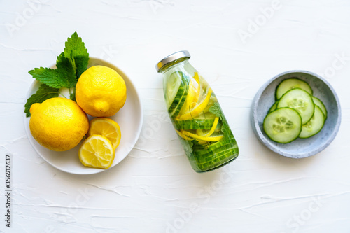 Eine Glasflasche mit Wasser, Zitronen und Gurken Scheiben, eine weiße Schüssel mit ganzen Zitronen auf einem weißen Hintergrund, Flat lay, Erfrischung, Wasser mit Geschmack